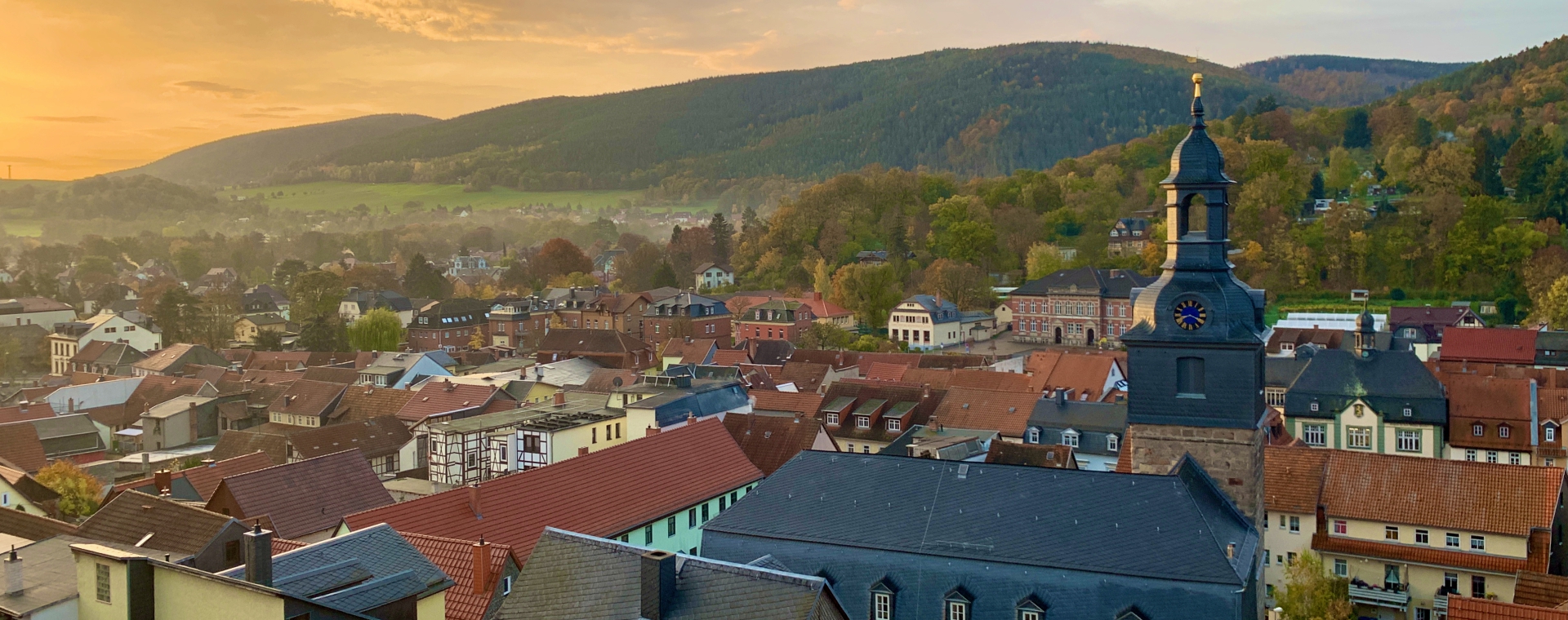 Blick auf eine malerische Stadt im Abendlicht, umgeben von sanften Hügeln. Die Dächer der Gebäude sind in verschiedenen Farben gehalten, während ein markanter Kirchturm im Vordergrund hervorsticht. Die Landschaft zeigt herbstliche Bäume und eine ruhige Atmosphäre. (Text KI generiert)