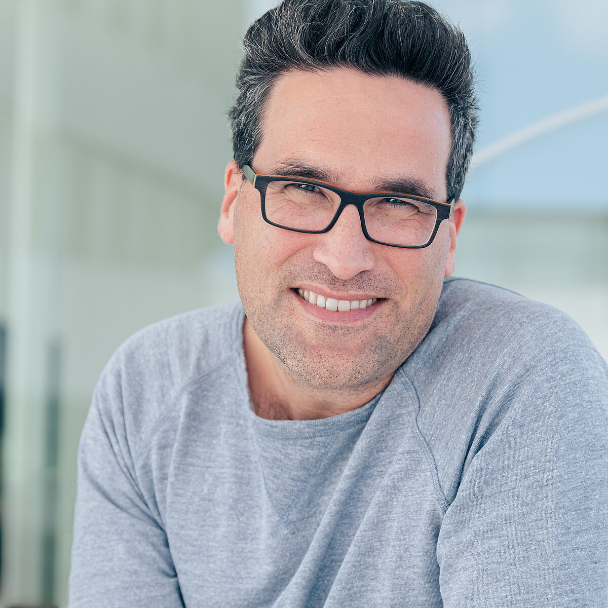 Ein lächelnder Mann mit Brille trägt ein graues Sweatshirt. Er sitzt in einem hellen, modernen Raum mit großen Fenstern im Hintergrund. (Text KI generiert)