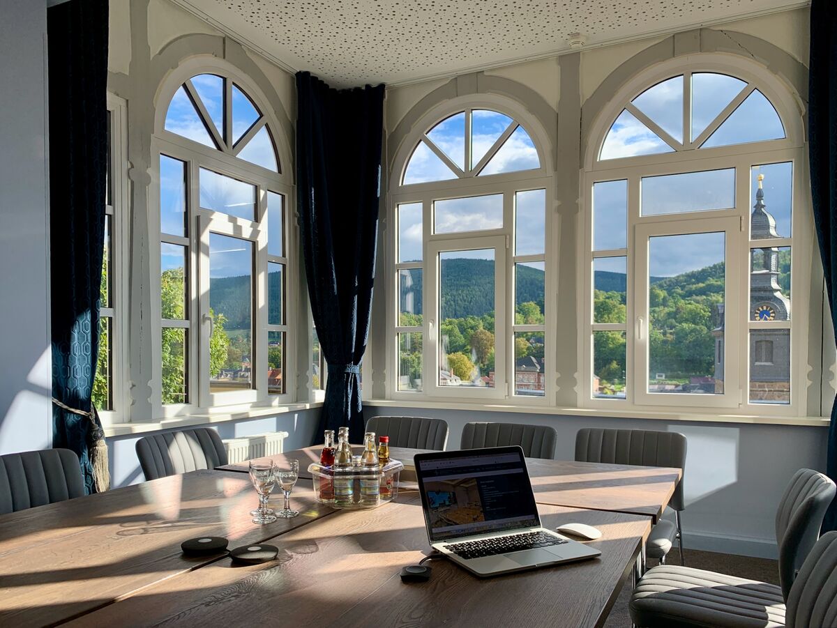 Ein heller Besprechungsraum mit großen Fenstern, die einen Blick auf grüne Hügel und Bäume bieten. Ein großer Tisch mit Stühlen ist in der Mitte, und ein Laptop steht auf dem Tisch. Die Wände sind hell und die Fensterrahmen sind architektonisch gestaltet. (Text KI generiert)