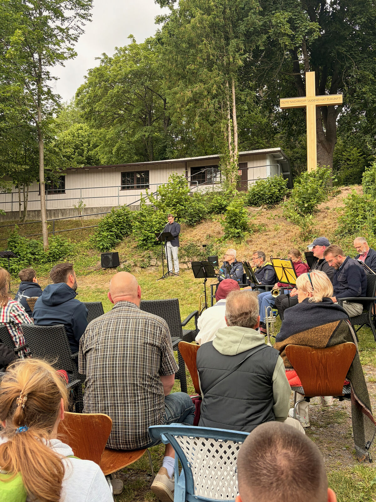 Eine Gruppe von Menschen sitzt auf Stühlen im Freien und hört einem Redner zu, der auf einer kleinen Bühne steht. Im Hintergrund ist ein großes Kreuz und ein Gebäude zu sehen, umgeben von Bäumen und Gras.