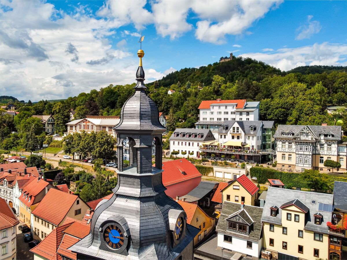 Blick auf eine Stadt mit historischen Gebäuden und einem markanten Kirchturm. Im Hintergrund sind grüne Hügel und ein Schloss sichtbar. Der Himmel ist blau mit einigen Wolken.