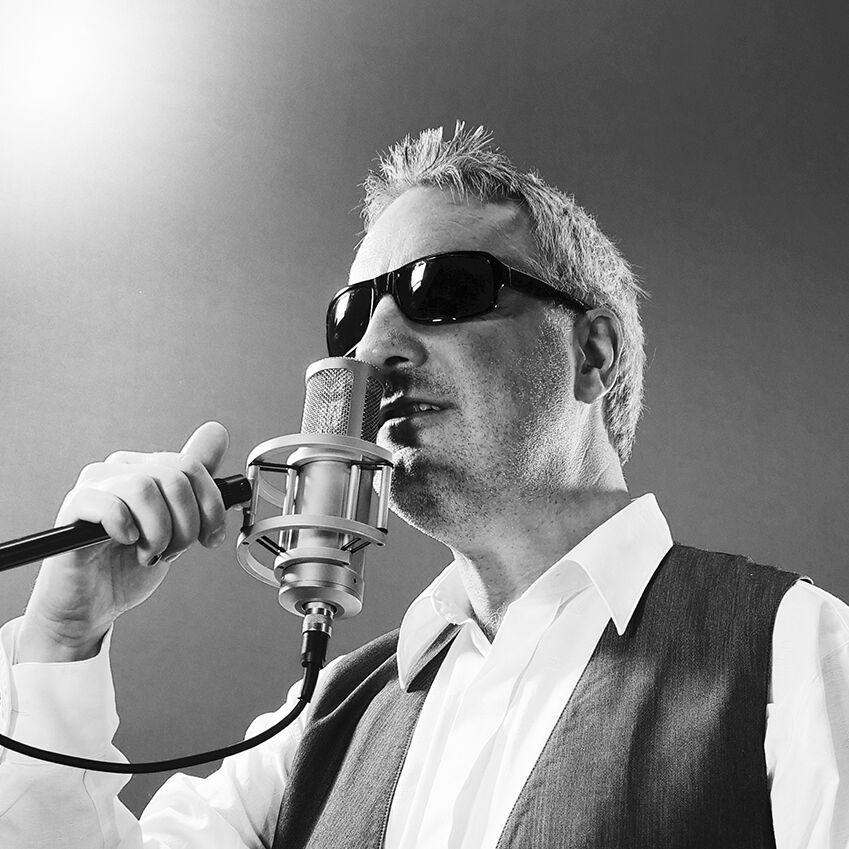 Ein Mann mit Sonnenbrille singt in ein Mikrofon. Er trägt eine Weste über einem weißen Hemd und hat graue Haare. Der Hintergrund ist neutral und unscharf, was den Fokus auf den Sänger lenkt.
