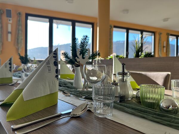 Ein gedeckter Tisch in einem Restaurant mit mehreren Gedecken, Servietten in Weiß und Grün, Gläsern und Besteck. Im Hintergrund sind große Fenster mit Blick auf eine Berglandschaft. Pflanzen und Dekorationen ergänzen die Tischgestaltung. (Text KI generiert)