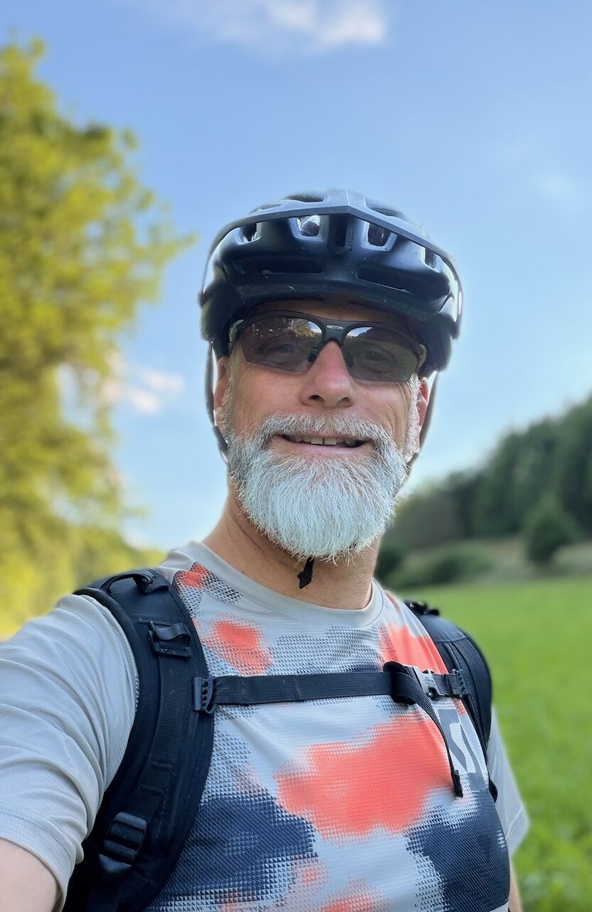Ein Mann mit grauem Bart und Brille trägt einen Fahrradhelm und ein sportliches T-Shirt. Er lächelt in die Kamera, während er auf einem Weg in der Natur steht, umgeben von grünen Wiesen und Bäumen unter einem blauen Himmel.