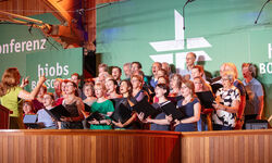 Ein gemischter Chor mit vielen Sängern steht auf einer Bühne und singt. Eine Dirigentin leitet den Chor. Im Hintergrund sind grüne Banner mit Texten zu sehen. Die Atmosphäre ist festlich und die Sänger sind konzentriert. (Text KI generiert)