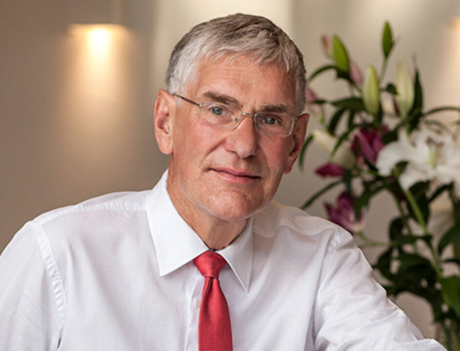 Ein älterer Mann mit grauen Haaren und Brille sitzt an einem Tisch. Er trägt ein weißes Hemd und eine rote Krawatte. Im Hintergrund sind Blumen zu sehen, die eine freundliche Atmosphäre schaffen.