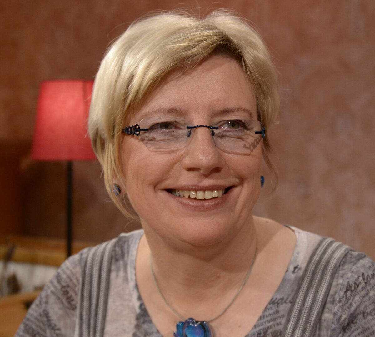 Eine lächelnde Frau mit blonden, kurzen Haaren und einer Brille sitzt in einem gemütlichen Raum. Im Hintergrund ist eine Lampe zu sehen. Sie trägt ein graues Oberteil mit einem Muster und einen blauen Anhänger.