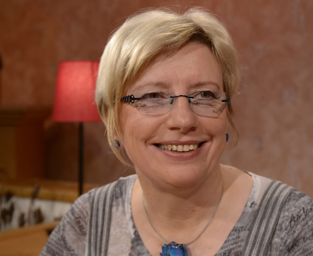 Eine lächelnde Frau mit blonden, kurzen Haaren und einer Brille sitzt in einem gemütlichen Raum. Im Hintergrund ist eine Lampe zu sehen. Sie trägt ein graues Oberteil mit einem Muster und einen blauen Anhänger.