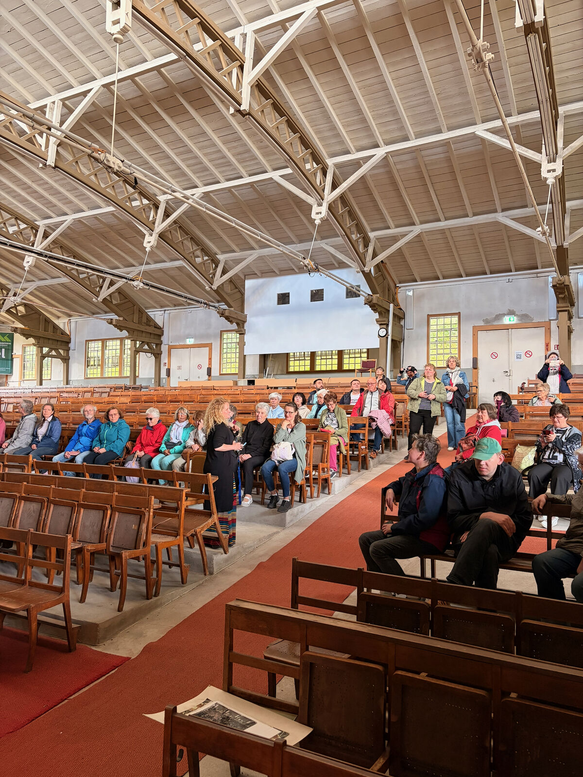 Eine Gruppe von Menschen sitzt und steht in einem großen, hellen Raum mit Holzbänken. Einige Personen unterhalten sich, während andere auf Stühlen sitzen. Die Architektur des Raumes zeigt hohe Decken und große Fenster.