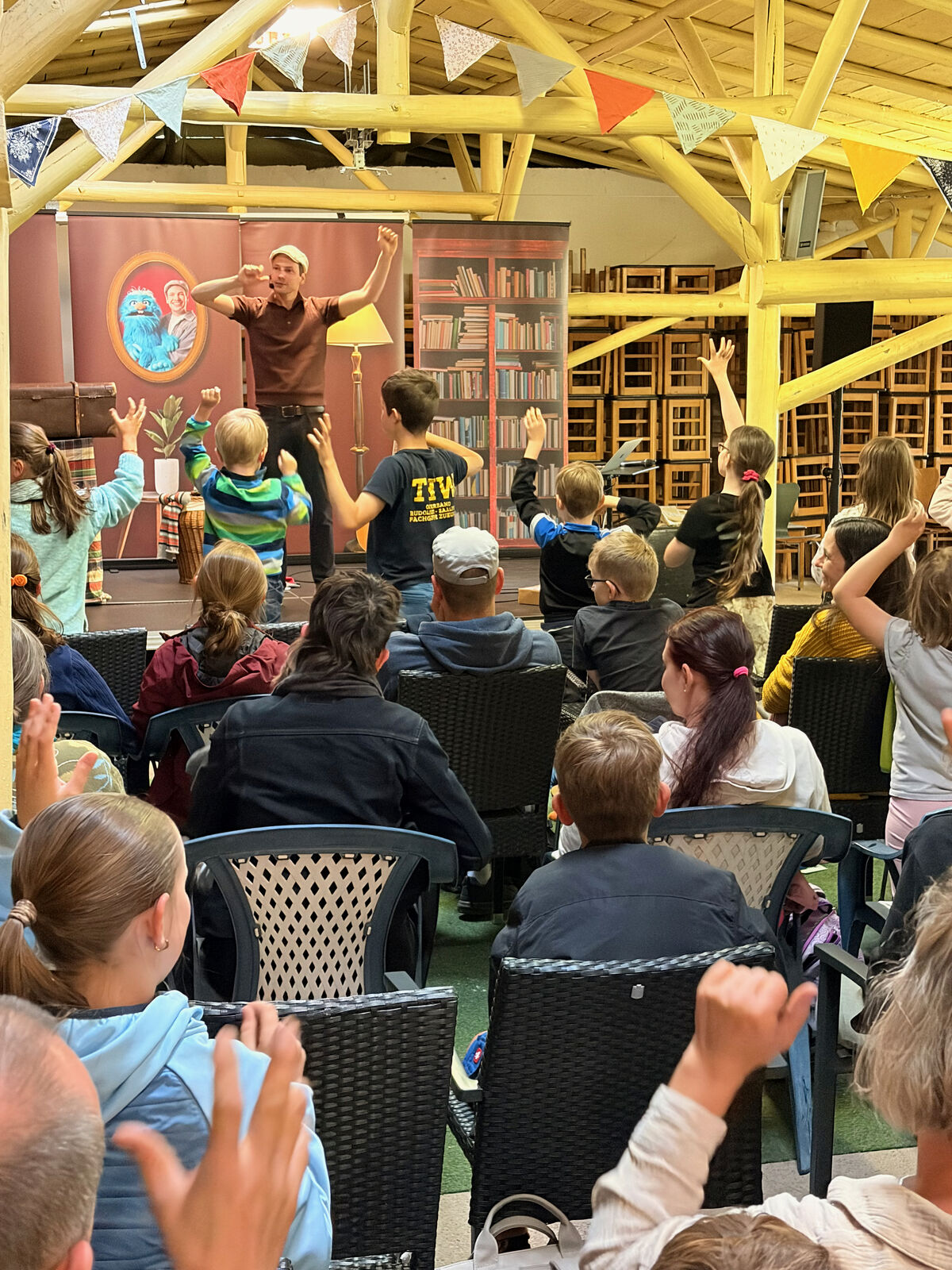 Eine Gruppe von Kindern und Erwachsenen sitzt auf Stühlen und schaut auf eine Bühne, auf der ein Performer steht. Der Performer gestikuliert mit den Armen, während die Zuschauer ihre Hände heben und aktiv teilnehmen. Im Hintergrund sind Bücherregale und eine bunte Dekoration zu sehen.