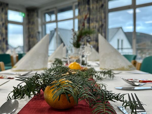 Ein festlich gedeckter Tisch mit weißen Servietten in Form von Hüten, einer roten Tischdecke und einer Dekoration aus Tannenzweigen und einer Orange in der Mitte. Im Hintergrund sind Fenster und Stühle sichtbar. (Text KI generiert)