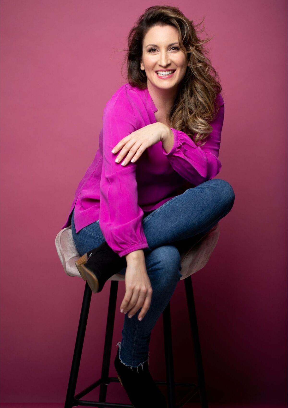 Eine lächelnde Frau sitzt auf einem Hocker, trägt ein pinkes Oberteil und Jeans. Sie hat lockige Haare und posiert entspannt, während sie einen Arm über das Knie legt. Der Hintergrund ist ebenfalls pink. (Text KI generiert)