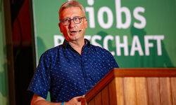 Ein Mann mit Brille und blauem Hemd spricht an einem Rednerpult. Im Hintergrund ist ein grünes Banner mit dem Text "Jobs" und "Wirtschaft" zu sehen. Die Szene vermittelt eine Atmosphäre einer Präsentation oder Konferenz.