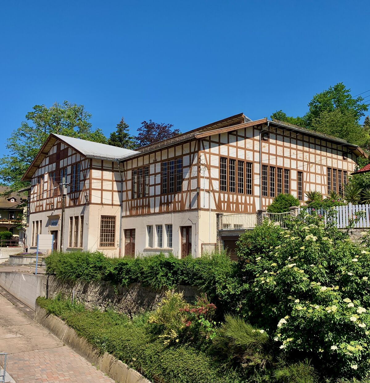 Ein Fachwerkhaus mit großen Fenstern und einem modernen Anbau steht in einer ruhigen Straße. Umgeben von Bäumen und Sträuchern, strahlt die Szene unter einem klaren blauen Himmel. (Text KI generiert)