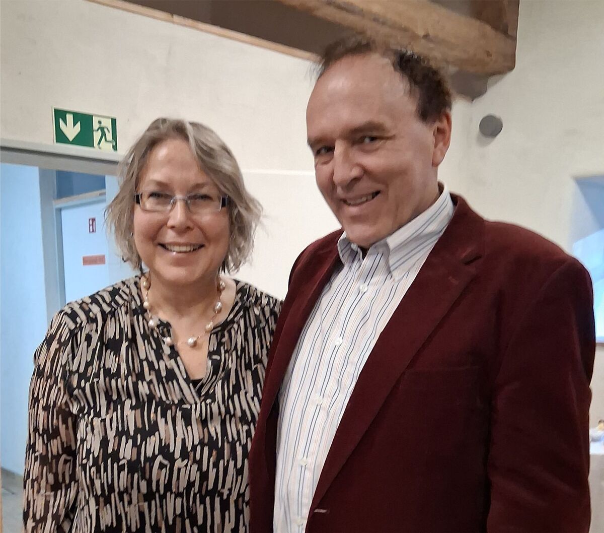 Zwei Personen stehen lächelnd nebeneinander in einem Innenraum. Die Frau trägt eine gestreifte Bluse und hat schulterlange, graue Haare. Der Mann trägt einen roten Blazer und ein gestreiftes Hemd. Im Hintergrund sind unscharfe Möbel und eine Notausgangszeichen zu sehen. (Text KI generiert)