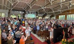 Eine große Menschenmenge steht in einem Gottesdienstraum, viele heben die Hände. Die Atmosphäre ist lebhaft und engagiert, mit Bänken und einer Bühne im Hintergrund. Helle Fenster lassen Tageslicht herein.