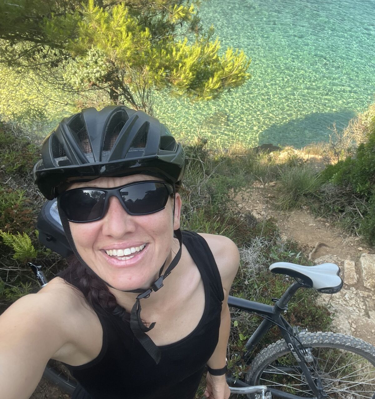 Eine Person mit Helm und Sonnenbrille lächelt in die Kamera, während sie auf einem schmalen Pfad neben einem klaren Gewässer steht. Im Hintergrund sind grüne Sträucher und Bäume zu sehen, sowie ein Fahrrad, das neben der Person steht. (Text KI generiert)