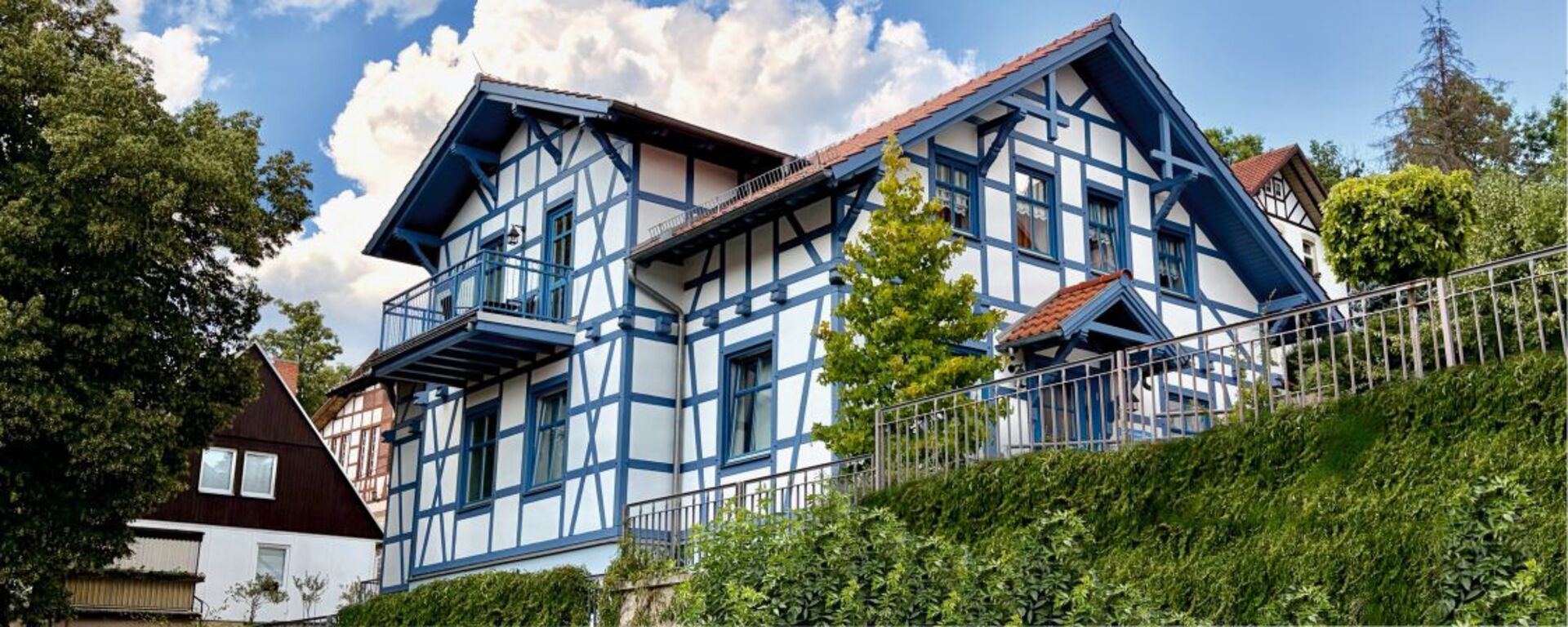 Ein zweigeschossiges, modernes Fachwerkhaus mit blauen und weißen Wänden sowie einem roten Satteldach. Es hat einen Balkon im oberen Stockwerk und ist von Bäumen und einem kleinen Garten umgeben. Der Himmel ist klar und sonnig.