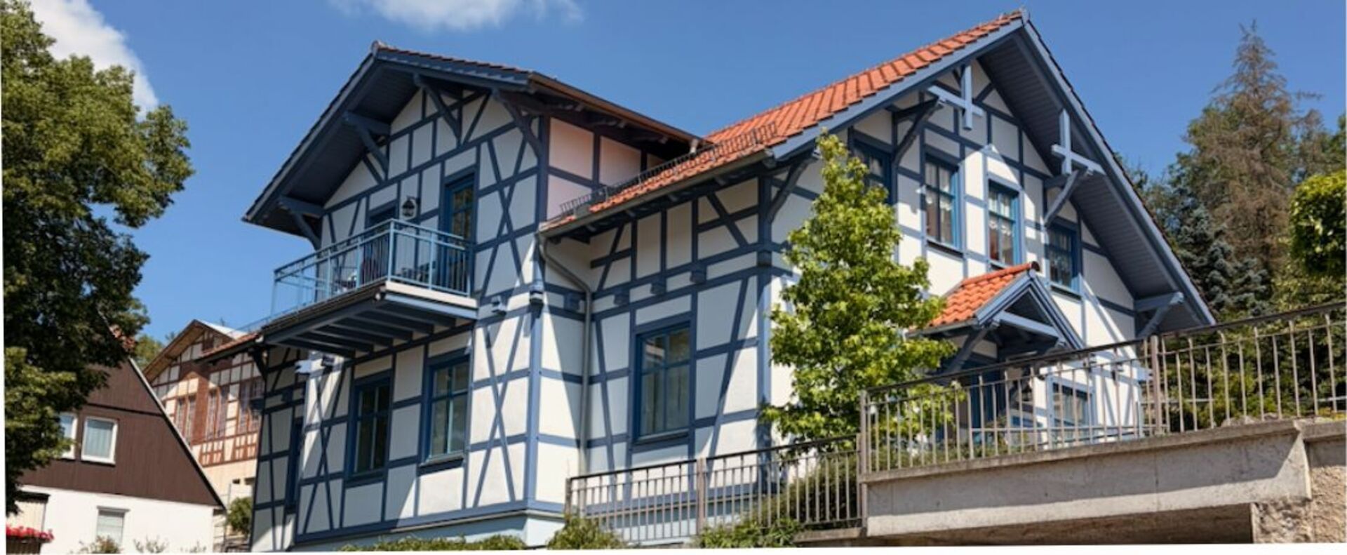 Ein zweigeschossiges, modernes Fachwerkhaus mit blauen und weißen Wänden sowie einem roten Satteldach. Es hat einen Balkon im oberen Stockwerk und ist von Bäumen und einem kleinen Garten umgeben. Der Himmel ist klar und sonnig.