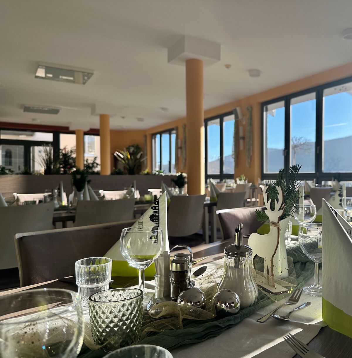 Ein eleganter Restaurantbereich mit Tischen, die mit weißen und grünen Servietten sowie Gläsern und Besteck gedeckt sind. Große Fenster lassen Tageslicht herein und bieten einen Blick auf die Landschaft. Pflanzen dekorieren den Raum. (Text KI generiert)