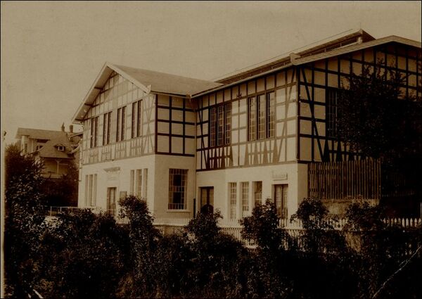 Ein historisches Gebäude mit Fachwerk-Architektur, das eine große Fensterfront hat. Es ist von Bäumen und Sträuchern umgeben. Die Fassade ist in hellen und dunklen Farben gehalten, was einen Kontrast zur Umgebung bildet. (Text KI generiert)