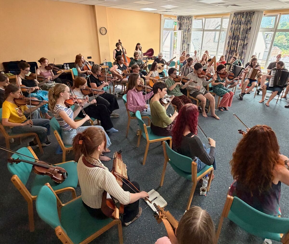 Eine Gruppe von etwa 40 Personen sitzt in einem Raum und spielt verschiedene Musikinstrumente, hauptsächlich Geigen. Einige Teilnehmer haben auch andere Instrumente wie Akkordeons. Der Raum ist hell erleuchtet und hat große Fenster mit Blick nach draußen.