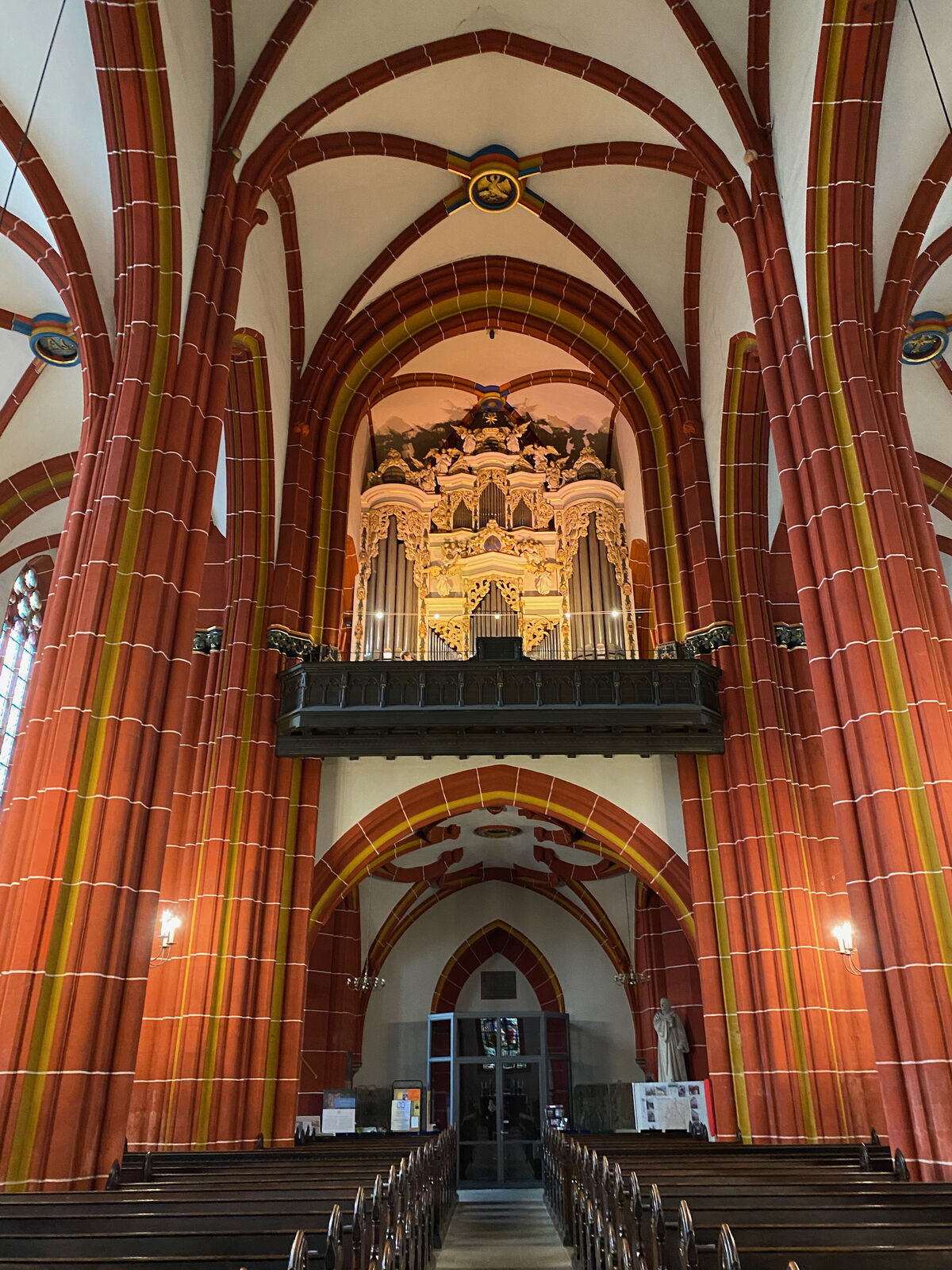 Innenansicht einer Kirche mit bunten, hohen Säulen und einem prächtigen Orgelbau im Hintergrund. Die Wände sind rot und gelb gestrichen, und die Bänke sind aufgereiht. Das Licht fällt sanft auf die Orgel und die Architektur. (Text KI generiert)