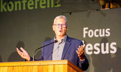 Ein Redner steht an einem Holzpult und spricht mit erhobenen Händen. Im Hintergrund sind die Worte "Focus Jesus" und "Konferenz" zu sehen. Der Redner trägt eine Brille und einen grauen Anzug.