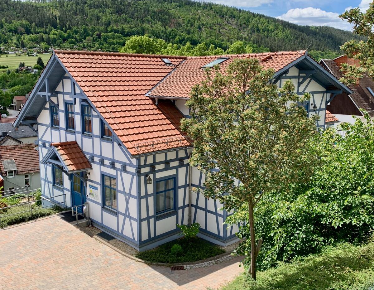 Ein zweigeschossiges Fachwerkhaus mit rotem Satteldach und weißen Wänden. Es steht auf einem leicht erhöhten Grundstück, umgeben von Bäumen und Sträuchern. Im Hintergrund sind sanfte Hügel und ein blauer Himmel mit einigen Wolken zu sehen.