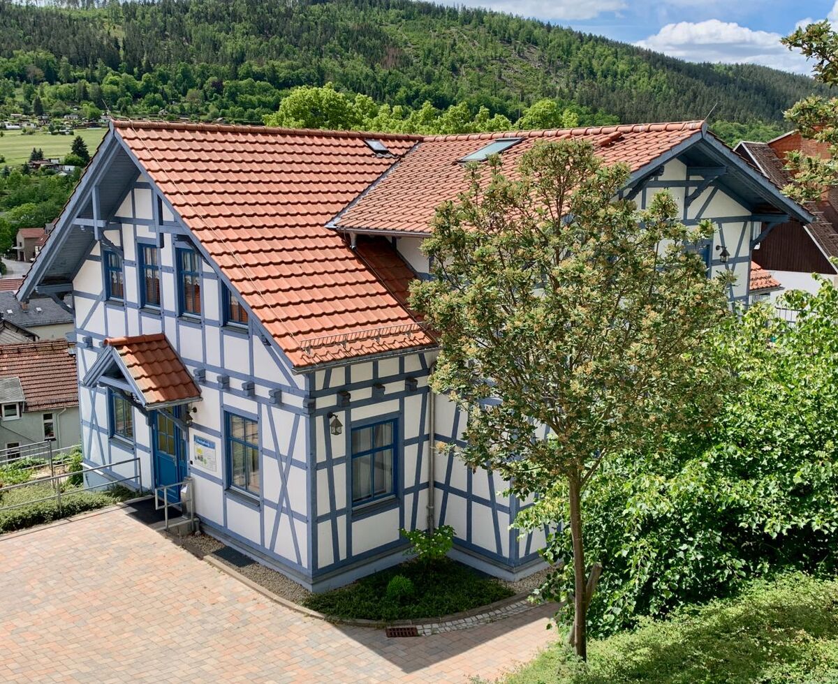 Ein zweigeschossiges Fachwerkhaus mit rotem Satteldach und weißen Wänden. Es steht auf einem leicht erhöhten Grundstück, umgeben von Bäumen und Sträuchern. Im Hintergrund sind sanfte Hügel und ein blauer Himmel mit einigen Wolken zu sehen.